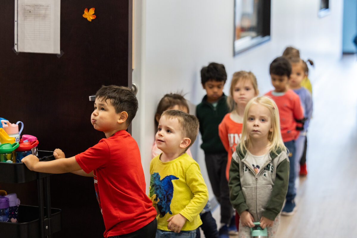 9 children are lined up and walking into their classroom. The child in the front of the line is pushing a cart filled with water bottles.