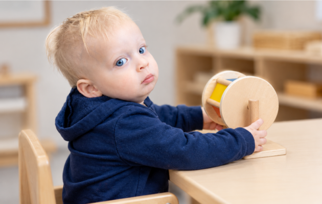 Picture depicts a two year old sitting at a table looking at the camera while holding a Montessori Spinning Drum.